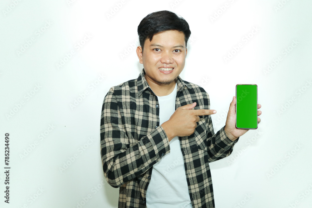 Young Asian man smiles happily while showing the green screen of his mobile phone and pointing at it Young Asian man smiles happily while showing the green screen of his mobile phone and pointing at it