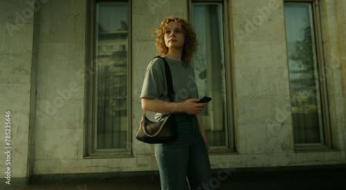 Confident young caucasian woman with curly hair standing in the city looking away holding mobile phone. Woman in casual outfit posing near part of old building.