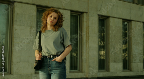 Positive woman with curly hairdressed in grey t-shirt posing against city building looking at camera. Copy space.
