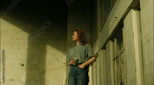 Friendly attractive young curly haired woman relaxing outside in an urban street, drinking takeaway coffee.