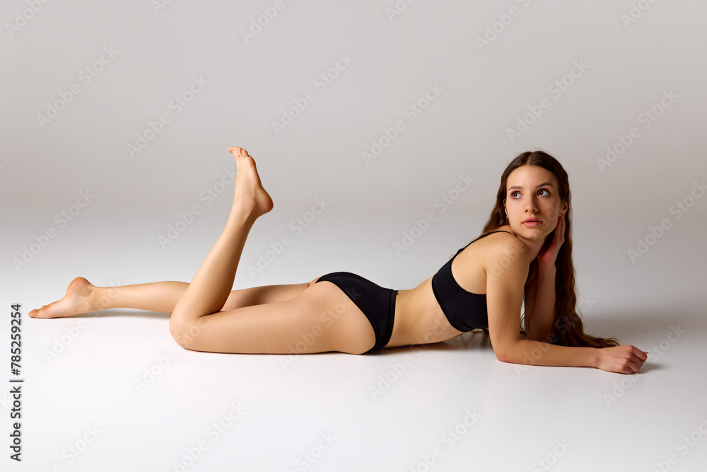 Beach season. Woman lying on floor and posing playfully in black swimwear against white studio background. Concept of beauty and health, depilation, anti-cellulite program, dieting.