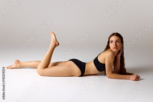Beach season. Woman lying on floor and posing playfully in black swimwear against white studio background. Concept of beauty and health, depilation, anti-cellulite program, dieting.