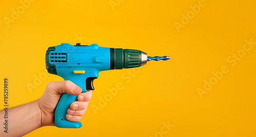 A blue power drill is being held by a person. The drill is pointed at the camera. The image has a bright yellow background. blue Cordless drill-driver in a hand photo