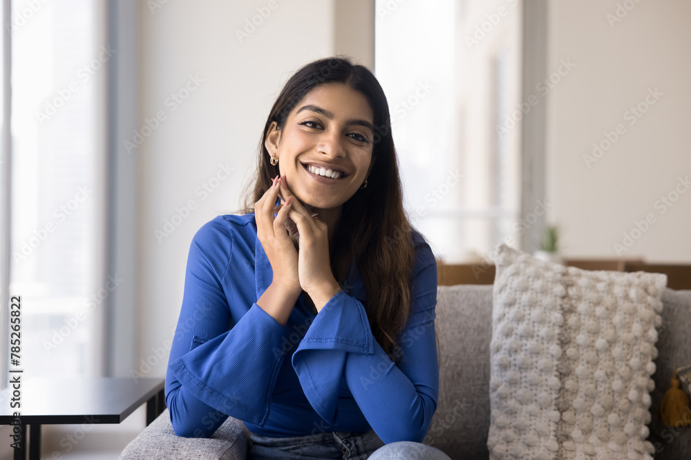 Positive beautiful young Indian woman touching face, facial skin, sitting on home couch, looking at camera with toothy smile. Happy relaxed beauty care female model posing for portrait Positive beautiful young Indian woman touching face, facial skin, sitting on home couch, looking at camera with toothy smile. Happy relaxed beauty care female model posing for portrait