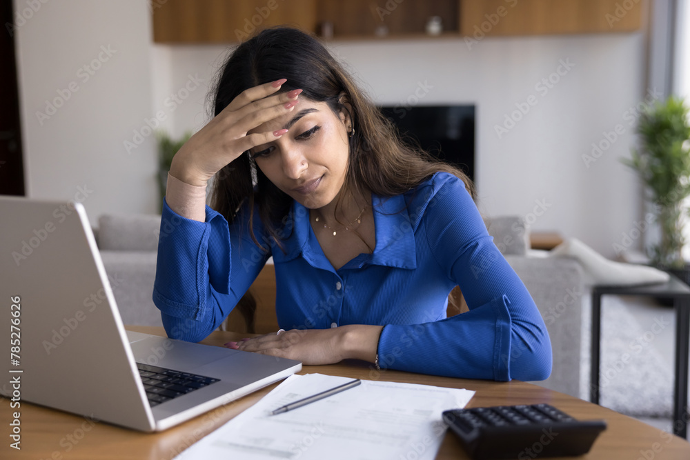 Worried sad young Indian homeowner woman thinking on money problems sitting at home workplace table with paper bills, invoices, calculator, laptop, touching head, looking at documents Worried sad young Indian homeowner woman thinking on money problems sitting at home workplace table with paper bills, invoices, calculator, laptop, touching head, looking at documents