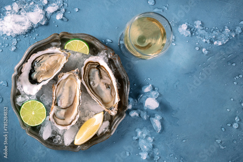 Oysters on a plate with lemon and lime, on ice, overhead flat lay shot on a blue background, with a glass of white wine, with copy space
