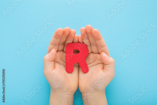 Baby boy hands holding red R letter on light blue table background. Pastel color. Time to learning. Closeup. Point of view shot. Top down view.