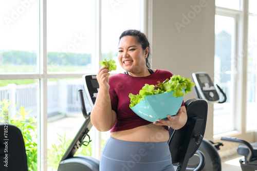 Overweight Asian female happy to eat organic green vegetable salad for healthy in gym. Weight loss workout, healthy lifestyle concept.