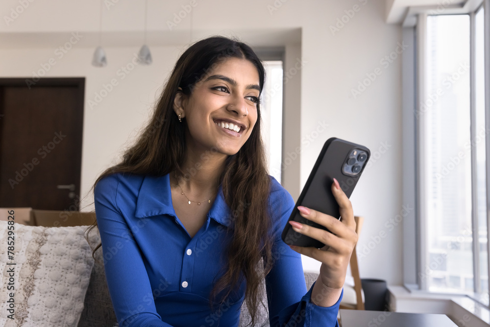 Happy dreamy young 20s Indian girl using smartphone on couch, holding gadget, looking away, smiling, thinking on Internet communication, smart home technology, laughing Happy dreamy young 20s Indian girl using smartphone on couch, holding gadget, looking away, smiling, thinking on Internet communication, smart home technology, laughing