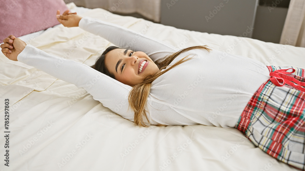A smiling young woman stretches comfortably on a bed in a bright bedroom, conveying a sense of leisure and relaxation. A smiling young woman stretches comfortably on a bed in a bright bedroom, conveying a sense of leisure and relaxation.