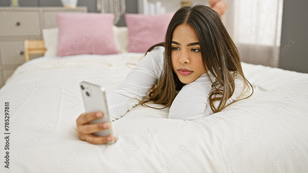 A young hispanic woman lies in bed holding a smartphone, portraying a relaxed indoor scene. A young hispanic woman lies in bed holding a smartphone, portraying a relaxed indoor scene.