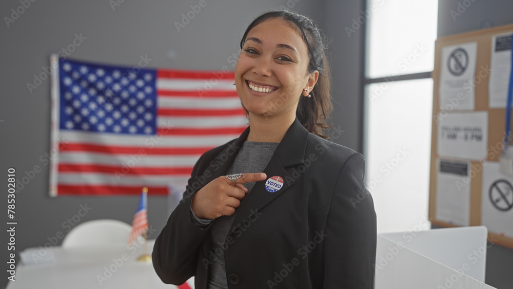 Fototapeta premium A smiling hispanic woman points to her 'i voted' sticker in a college voting center with an american flag backdrop