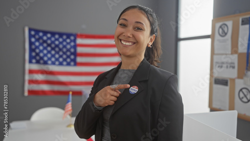A smiling hispanic woman points to her 'i voted' sticker in a college voting center with an american flag backdrop © Krakenimages.com