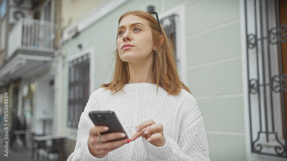 Caucasian woman with red hair in white sweater using smartphone on city street, looking contemplative.