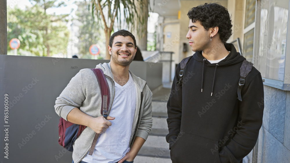 Two young men friends smiling and conversing on an urban city street with one carrying a backpack. Two young men friends smiling and conversing on an urban city street with one carrying a backpack.
