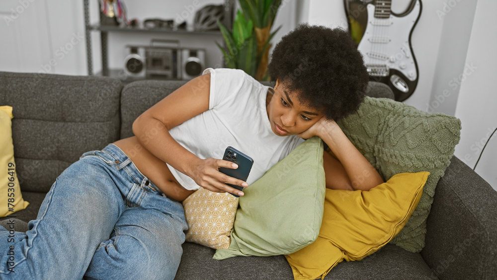 African american woman using smartphone on couch indoors with guitar in the background - obrazy, fototapety, plakaty 