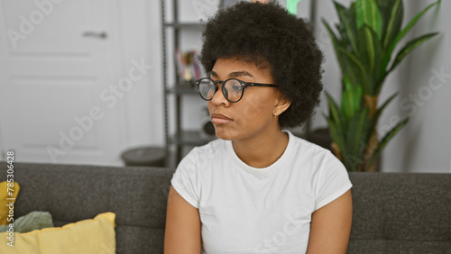 Thoughtful african american woman with curly hair wearing glasses, sitting on a couch indoors.