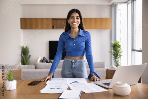 Happy ambitious Indian startup leader girl standing for portrait at home workplace table with marketing financial repots, laptop computer, looking at camera with toothy smile
