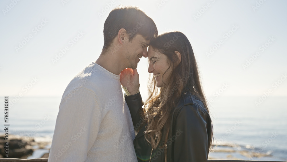 Close-up of a smiling couple caressing noses at a sunny seaside, displaying love and companionship with the ocean in the backdrop