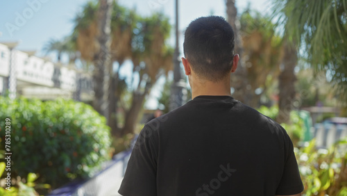A young hispanic man with a short haircut stands outdoors surrounded by greenery and palm trees in a sunny park.