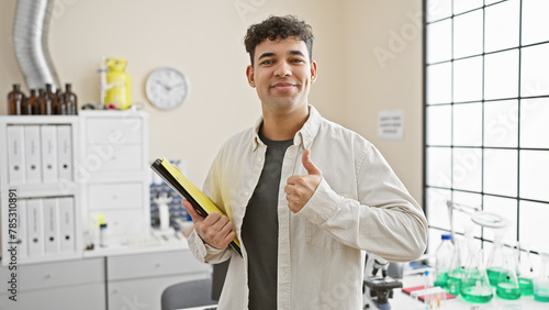 A confident young man with a folder gives a thumbs up in a modern laboratory setting, embodying professionalism and a positive attitude.