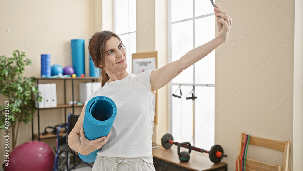 A middle-aged woman takes a selfie while holding a yoga mat in a well-equipped physiotherapy center, showcasing an active lifestyle. A middle-aged woman takes a selfie while holding a yoga mat in a well-equipped physiotherapy center, showcasing an active lifestyle.