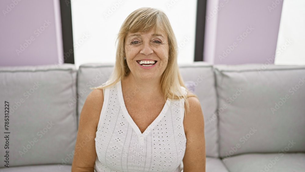 Confident middle age blonde woman joyfully resting, smiling beautifully on cozy sofa at home, radiating positivity.