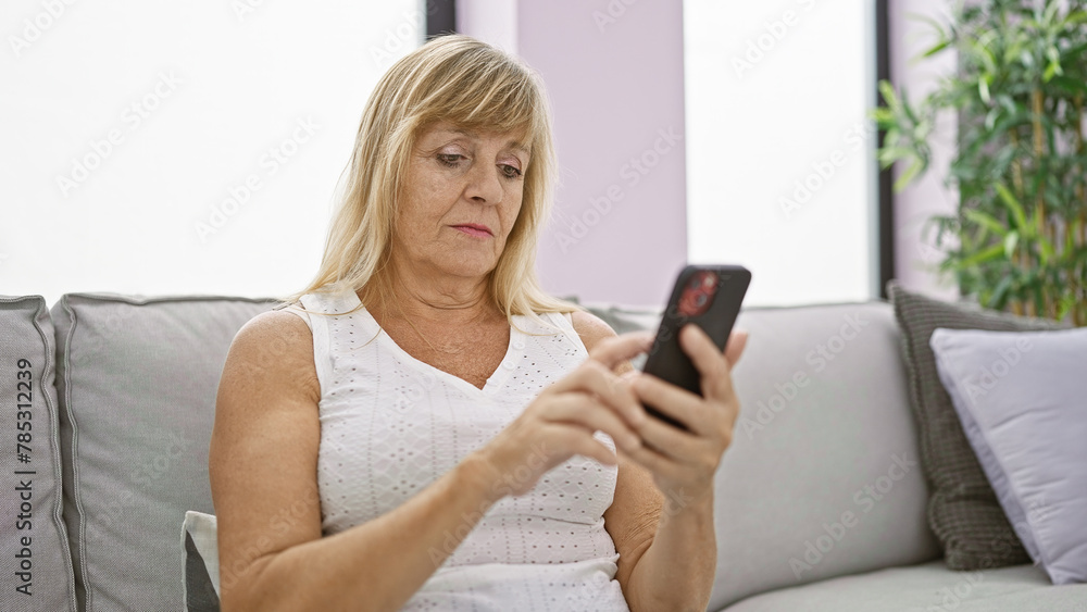 Serious mid-age blonde woman struggles with technology, upset expression as she sits on sofa indoors, typing message on smartphone, engrossed in the problem at hand in her stylish living room Serious mid-age blonde woman struggles with technology, upset expression as she sits on sofa indoors, typing message on smartphone, engrossed in the problem at hand in her stylish living room