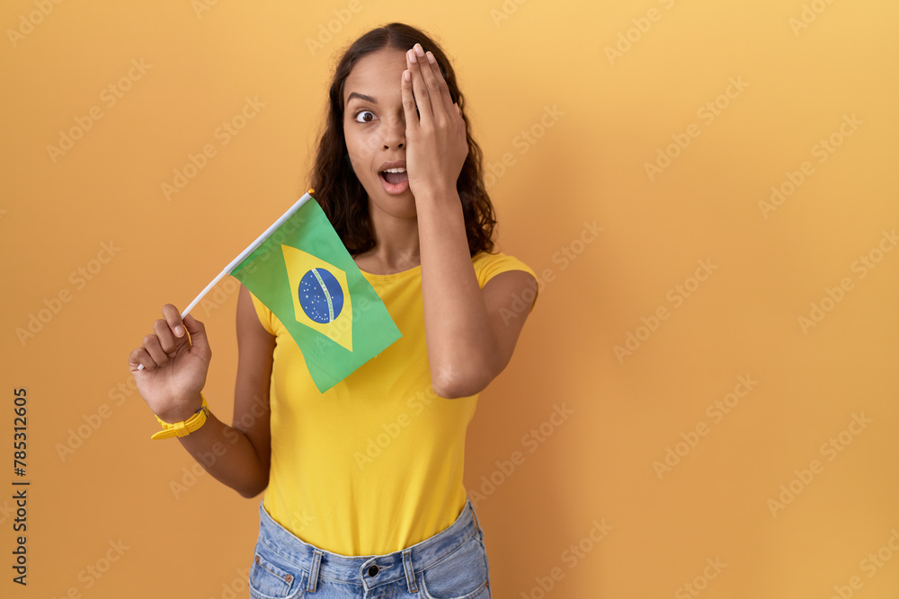 Young hispanic woman holding brazil flag covering one eye with hand, confident smile on face and surprise emotion. Young hispanic woman holding brazil flag covering one eye with hand, confident smile on face and surprise emotion.