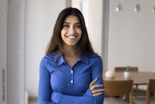 Happy confident 20s Indian girl home head shot. Positive beautiful young woman in casual posing for portrait in apartment, keeping arms crossed, looking at camera with toothy smile