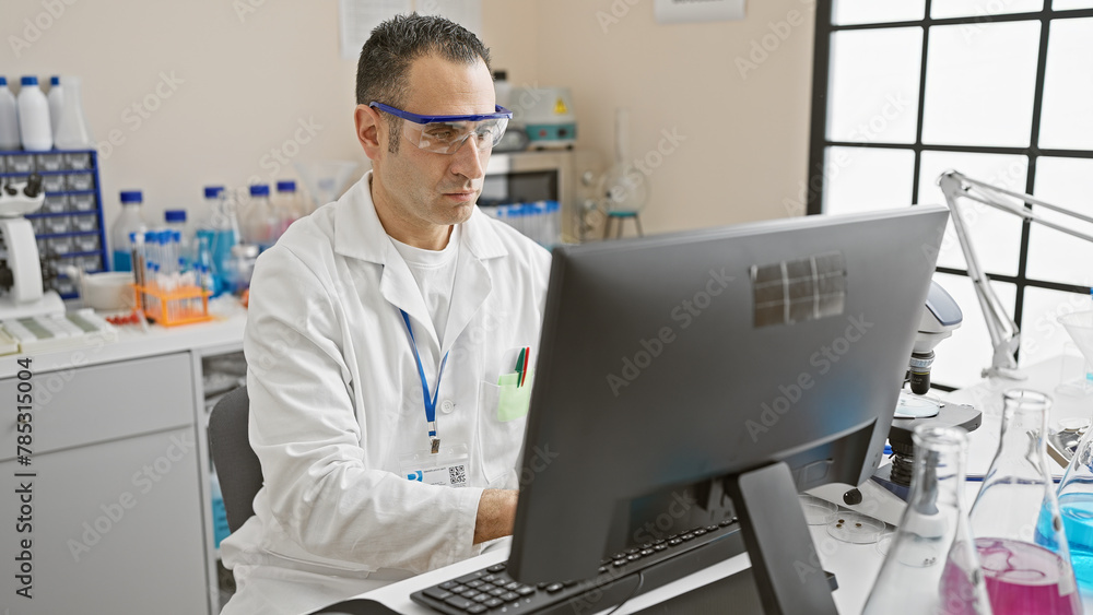 Hispanic scientist man in lab coat working at computer in modern laboratory indoor setting. Hispanic scientist man in lab coat working at computer in modern laboratory indoor setting.