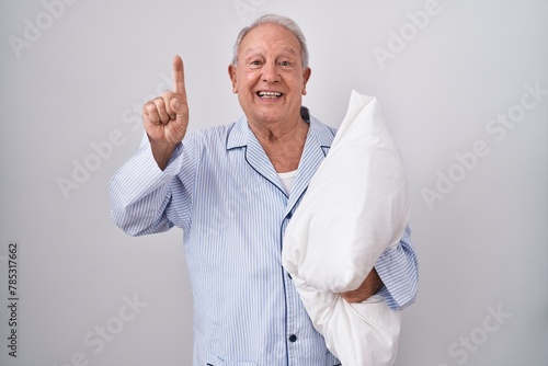 Senior man with grey hair wearing pijama hugging pillow smiling amazed and surprised and pointing up with fingers and raised arms.