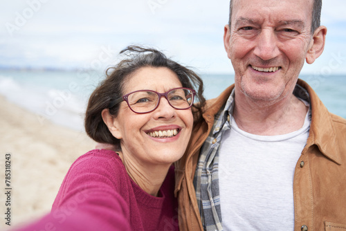Mature couple taking selfie with smartphone in the beach by the ocean in summer morning