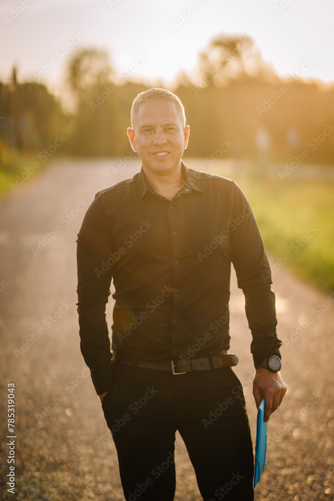 Obraz premium Valmiera, Latvia - August 17, 2024 - A man stands on a country road at sunset, holding a folder, with a pleasant smile on his face.