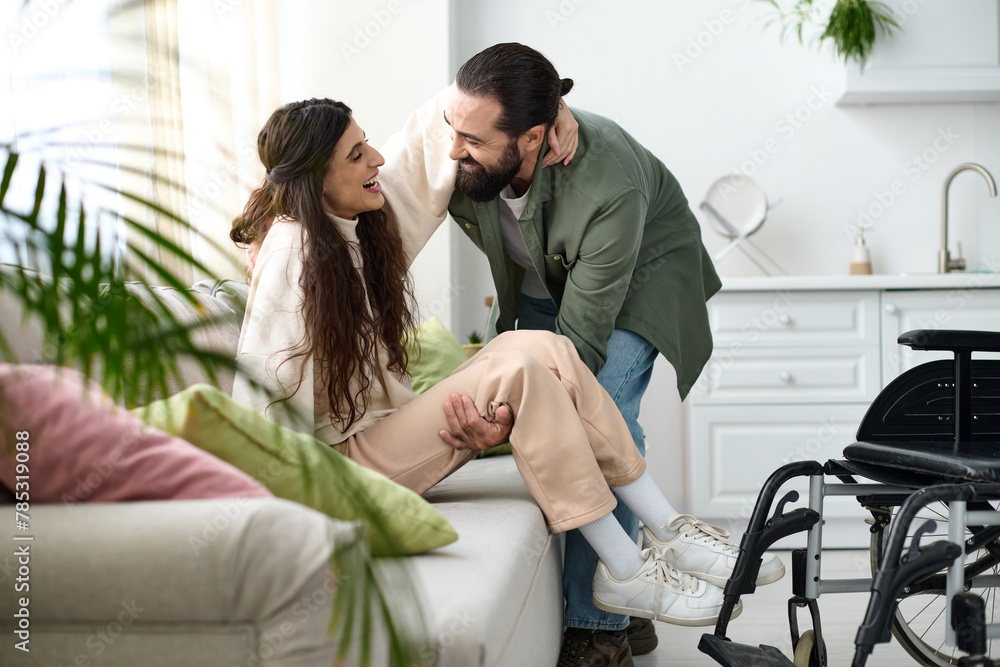 Obraz premium loving man in homewear helping his wife with mobility disability to get to couch from wheelchair