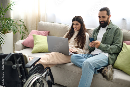 beautiful woman with mobility disability sitting on sofa with laptop next to her husband with phone