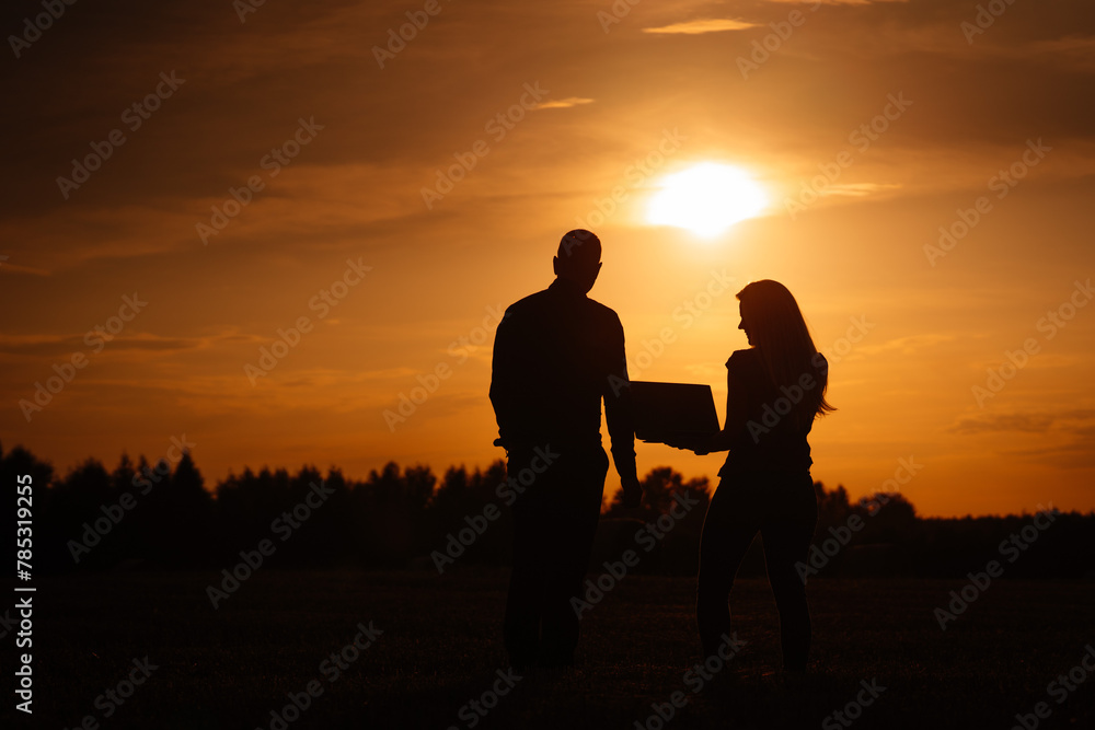 Valmiera, Latvia - August 17, 2024 - Silhouettes of a man and a woman with a laptop in a field during a vibrant sunset.