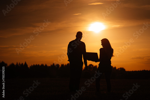 Valmiera, Latvia - August 17, 2024 - Silhouettes of a man and a woman with a laptop in a field during a vibrant sunset.