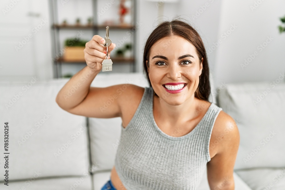 Young beautiful hispanic woman holding key of new house sitting on sofa at home