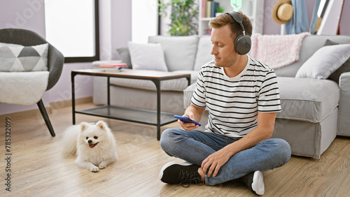 Engrossed young caucasian man sitting on the floor at home, deeply focused on a song playing on his smartphone using headphones while his adult dog patiently waits beside him