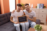 Homosexual gay couple holding blackboard with first home text winking looking at the camera with sexy expression, cheerful and happy face.
