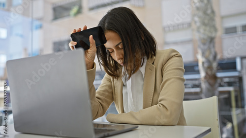 Bilde på lerret A stressed young hispanic woman at a desk with a laptop holding her phone to her temple in a beige office environment