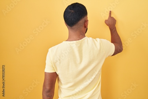 Young hispanic man standing over yellow background posing backwards pointing ahead with finger hand photo