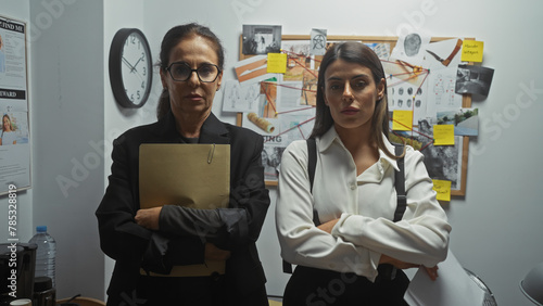 Two serious women detectives stand with crossed arms in a police department investigation room, surrounded by evidence.