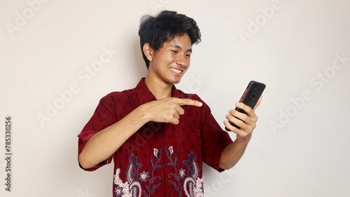 happy young handsome Asian man in batik shirt posing pointing at smartphone