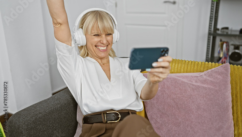 Cheerful mature woman with blonde hair enjoying music on headphones and taking a selfie at home. photo