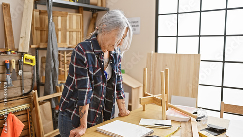 Serious-faced, grey-haired middle age woman, a skilled carpenter, standing amidst her carpentry workshop