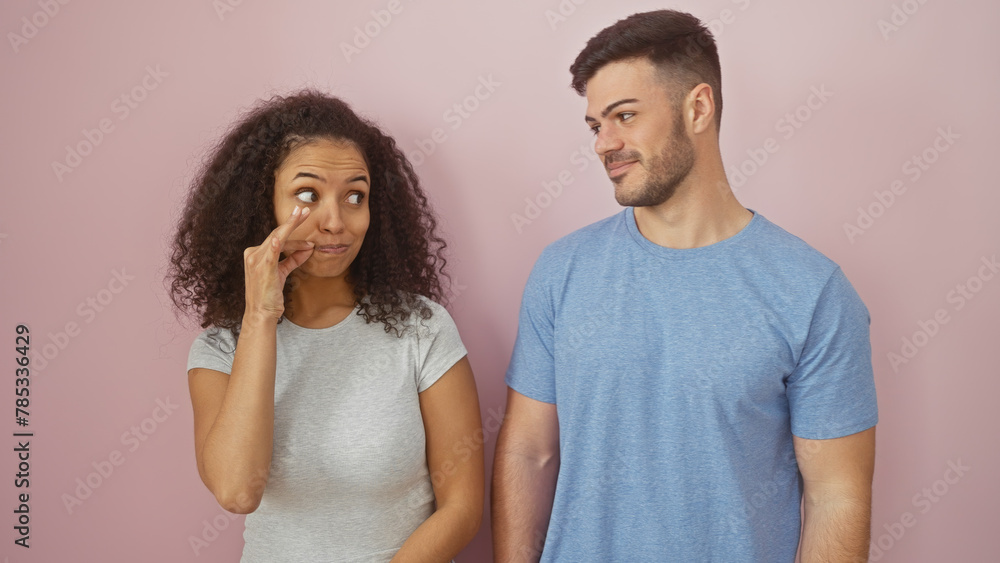 A woman gestures to silence herself beside a smiling man, both standing against a plain pink background. A woman gestures to silence herself beside a smiling man, both standing against a plain pink background.