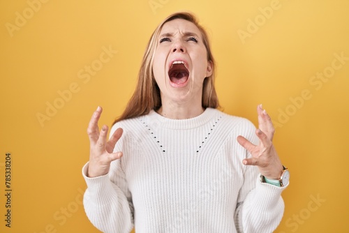 Quadro em tela Young caucasian woman wearing white sweater over yellow background crazy and mad shouting and yelling with aggressive expression and arms raised