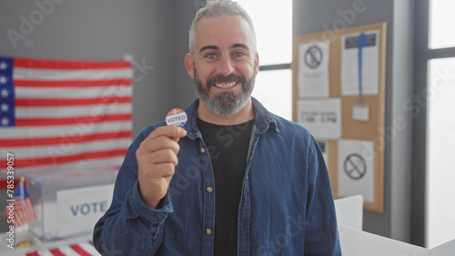 Smiling bearded man showing 'i voted' sticker with american flag and voting booths in background. photo